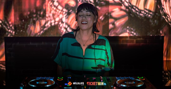 A DJ mid-laugh behind the decks, a huge projection filling the wall behind her