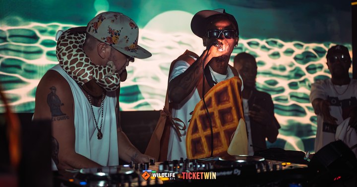 Two DJs side by side at the booth against a green and white honeycomb projection
