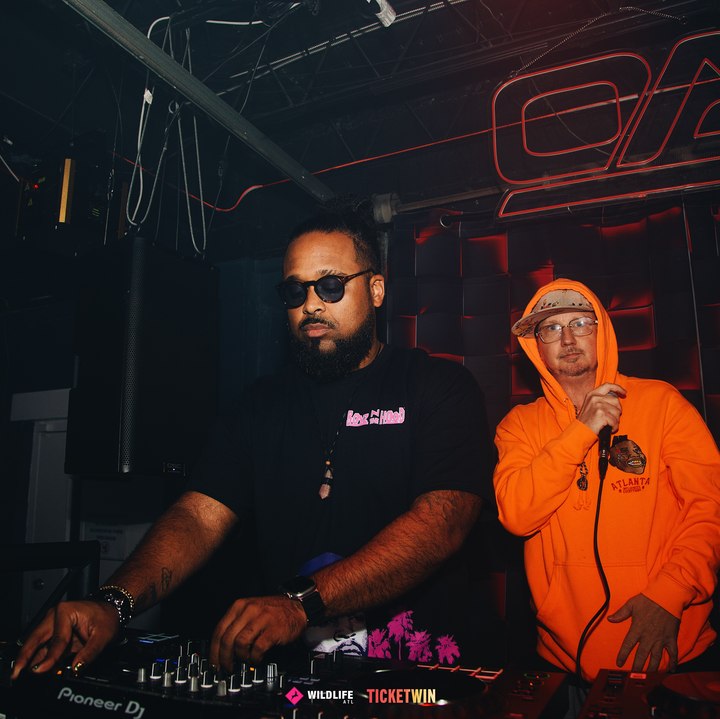 Two DJs side by side behind the decks, one on the mic in a bright orange hoodie