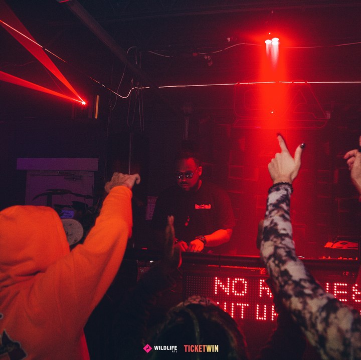 Hands up in the crowd under a red beam, the NO REQUESTS SHUT UP AND DANCE sign glowing behind the booth