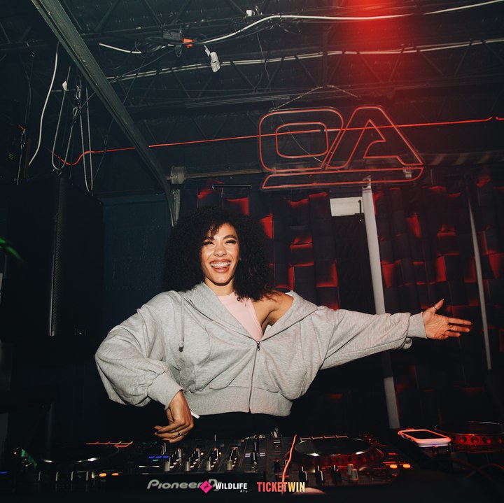 A DJ laughing with both arms flung wide behind the decks, the venue's neon monogram above her