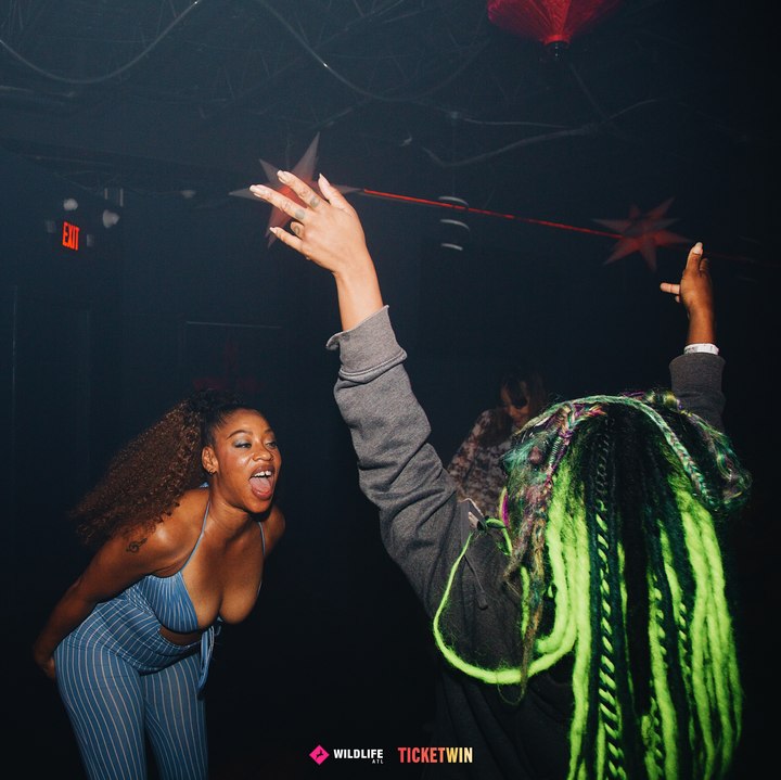 Two dancers mid-move, one with floor-length neon green locs, an arm thrown up between them