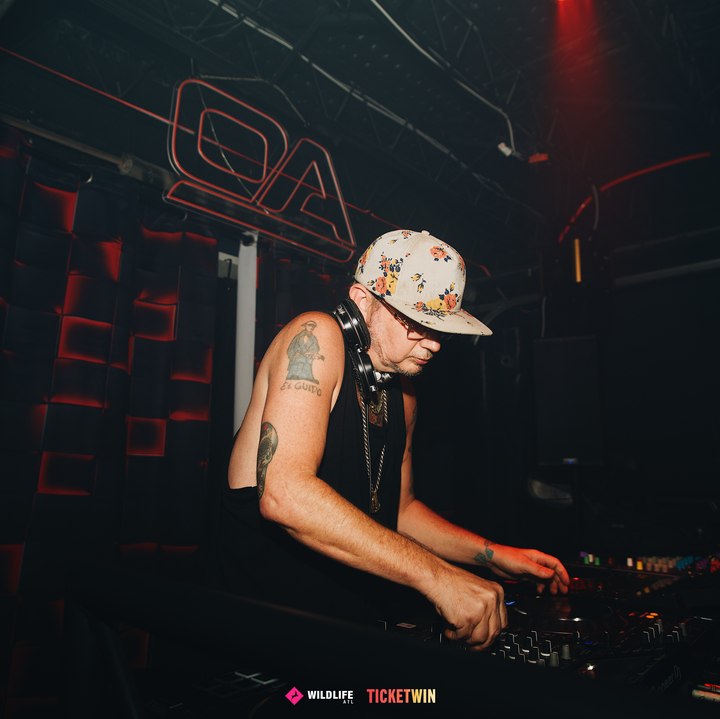 A DJ in a floral cap working the mixer with both hands under the neon monogram